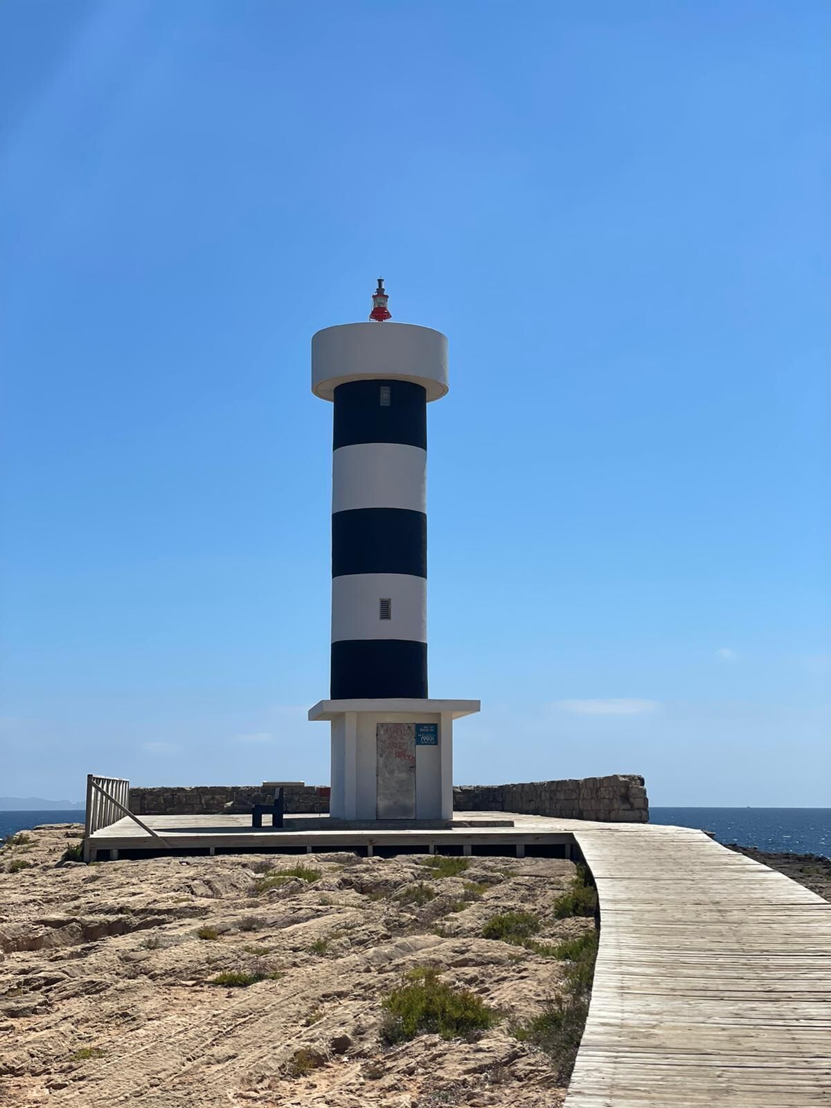 Ein Leuchtturm mit abwechselnd schwarzen und weißen Streifen, ein hölzerner Weg dorthin und felsiges Gelände im Vordergrund.