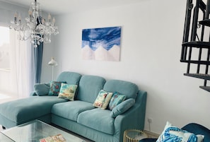 A teal sofa with decorative pillows, a glass coffee table, a chandelier, a wall-mounted painting, and a staircase.