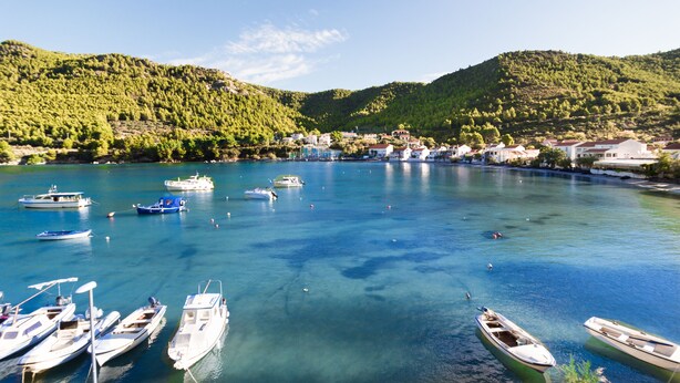 Un pueblo costero con barcos amarrados a la orilla, rodeado de colinas y agua azul cristalina.