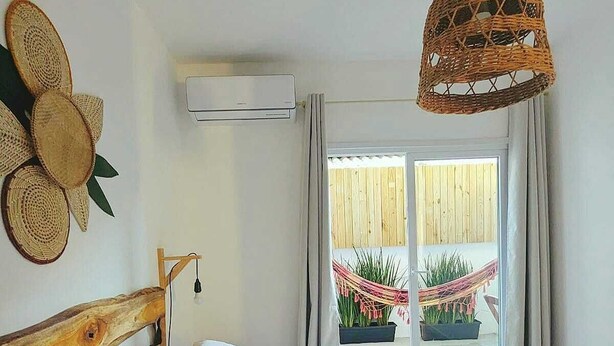 Une chambre à coucher avec une tête de lit en bois, un lit recouvert de draps blancs, un luminaire suspendu en fibres tressées et une vue sur une clôture en bois et des plantes à l’extérieur.
