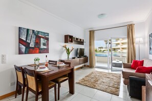 Relax in this stylish living room with a flat-screen TV, dining table, and sofa, offering a view of the pool and surrounding buildings from the balcony.