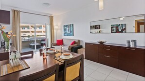 Moderni hotellihuone, jossa on ruokailutila, sohva, suuri ikkuna, josta on näkymä muihin hotellihuoneisiin, peili ja taulu seinällä.