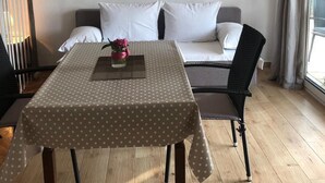 A hotel room with a bed, a dining table with a polka dot tablecloth, a painting of a sunset over the ocean on the wall, and a window with curtains. - Grapevine Studio Apartment (Zdrelac)