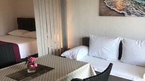 A hotel room with a bed, a sofa, a dining table with a vase of flowers, and a painting of a sunset over the ocean. - Grapevine Studio Apartment (Zdrelac)