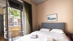A hotel room with a bed, a small table, a chair, a window with curtains, and a framed picture on the wall. - Ideal 4 bdr Apartment in Plaka for 10! (Athens)