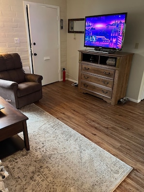 Una sala de estar con un sofá de cuero marrón, un mueble de madera con cajones, un televisor encima, una alfombra estampada y piso de madera.
