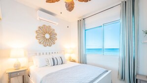 Een slaapkamer met een bed, een decoratief wandobject, een airconditioningapparaat, een nachtkastje en uitzicht op zee door een raam.