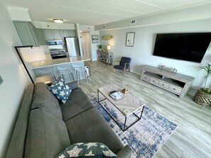 A modern living room with a grey sofa, a coffee table, and a TV. There is a kitchen area with a fridge and a dining table.
