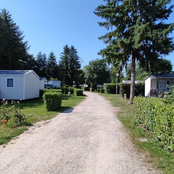 Un chemin de gravier traverse un terrain de camping où se trouvent des mobil-homes et une végétation luxuriante.