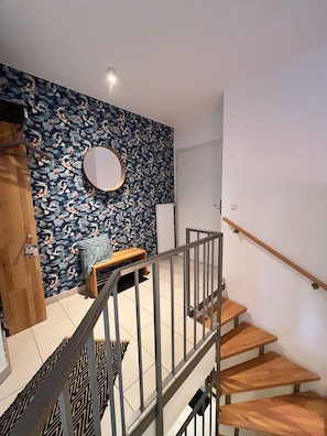 Un escalier avec des marches en bois et des rampes en métal, menant à un palier orné d’un papier peint à motifs floraux et agrémenté d’un miroir rond.