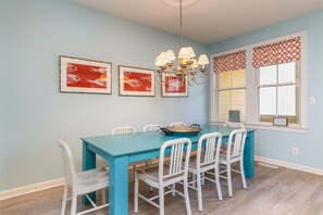 A dining area with a turquoise table and white chairs, framed artwork on the wall, and a window with patterned valance.