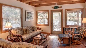 Ein gemütliches Wohnzimmer mit Holzbalkendecke, einer Couch, zwei Sesseln, einem Couchtisch und einem Bücherregal.