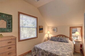 A bedroom with a bed, two bedside tables with lamps, a dresser, a window with blinds, and a decorative mirror. - 1214 - Toad Hall - Cozy Corolla Cottage | Walk to Beach & Village (Corolla)
