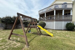 A wooden swing set with a yellow slide in a backyard with a white fence and a two-story building in the background. - 1418 - Windswept - Walk to Beach | Pool, Hot Tub & Wraparound Deck (Corolla)