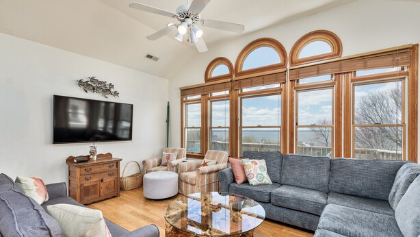 A spacious living room with a large flat-screen TV, a ceiling fan, and a view of the outdoors through large windows. - 1559 - Sound Retreat - Spacious Corolla Soundfront w/ Sunset Views & Pool (Corolla)
