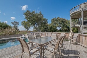 A wooden deck with a glass-top table and chairs, a pool, and a two-story building with a balcony.