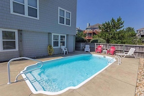 A rectangular swimming pool with a white edge, surrounded by a concrete deck. There are white and red lounge chairs, and a wooden fence in the background. - 1610 - Wave Hello - Walk to Beach | Private Pool & Hot Tub in Corolla (Corolla)