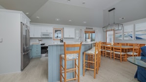 A modern kitchen with white cabinets, a central island, wooden chairs, and a dining area with a glass table. - 1618 - Dune Buggy - Walk to Beach | Private Pool & Hot Tub (Corolla)