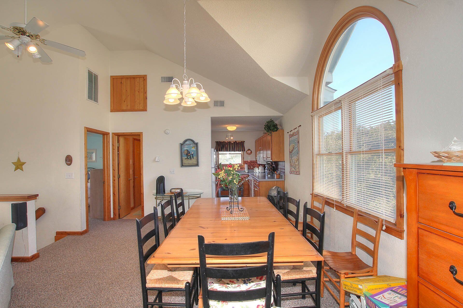 A spacious dining area with a long wooden table, black chairs, and a chandelier. There is a kitchen visible in the background with wooden cabinets and a window with blinds.