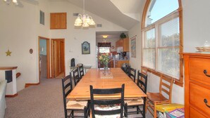 A spacious dining area with a long wooden table, black chairs, and a chandelier. There is a kitchen visible in the background with wooden cabinets and a window with blinds. - 1758 - Chesapeake Colony Escape - Dog-Friendly Corolla Retreat | Pool & Hot Tub (Corolla)