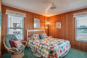 Ein gemütliches Schlafzimmer mit Holzwänden, einem gerafften Fensterbordu, einem Korbstuhl, einem Bett mit farbenfrohem Punkte-Überwurf und einer Nachttischlampe.