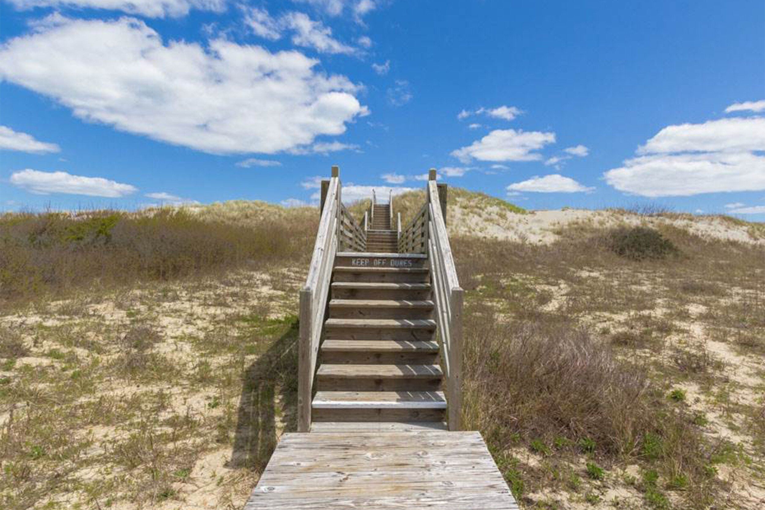 Eine Holztreppe führt zu einem Strandabschnitt unter blauem Himmel.