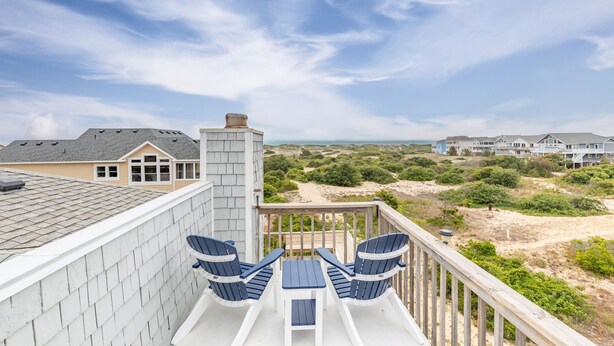 Ein Balkon mit zwei Adirondack-Stühlen und einem kleinen Tisch mit Blick auf einen Strand und weitere Gebäude.