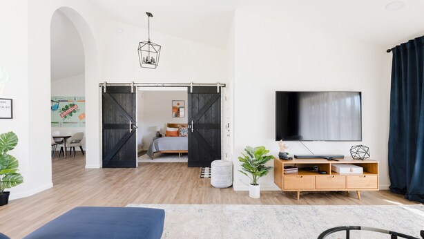 A modern living room with a large TV, a sofa, and a dining area visible through an open door. - Designer 4BR w/ Free Heated Pool & Fire Pit,  Hiking Trail Right Off the Yard (Scottsdale)