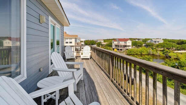 Une terrasse en bois avec des chaises Adirondack blanches, une balustrade et une vue sur une rivière et des maisons en arrière-plan.