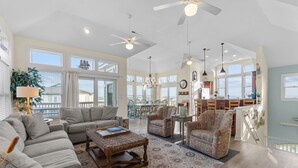 A spacious living room with a high ceiling, large windows, and a view of the ocean. There is a dining area with a table and chairs, and a kitchen in the background. - 2820 - Dabbling Duck - Spacious Duck Getaway | Pool, Hot Tub & More (Duck)