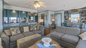 A living room with a sofa, armchair, and coffee table. There is a ceiling fan, a window with a view, and a bookshelf. - 2885 - Cobb Station, Oceanside home perched high on a rise with great ocean views & sea breezes (Duck)