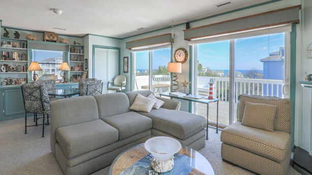 A living room with a gray sectional sofa, patterned chairs, a glass coffee table, and a view of the ocean through large windows. - 2885 - Cobb Station, Oceanside home perched high on a rise with great ocean views & sea breezes (Duck)