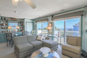 A living room with a gray sectional sofa, patterned chairs, a glass coffee table, and a view of the ocean through large windows. - 2885 - Cobb Station, Oceanside home perched high on a rise with great ocean views & sea breezes (Duck)