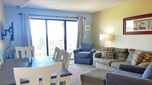 A living room with a dining table, two chairs, a sofa, and a couch. There is a window with curtains, a lamp, and a framed picture on the wall. - 2948 - Duck Nest, Perfect for smaller families or couples with unparalleled ocean views & amenities (Duck)