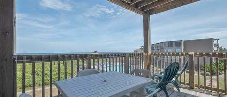 A covered outdoor area with a table and chairs, overlooking a beach and a pool.
