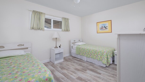 A bedroom with two beds, a window with curtains, a ceiling fan, a bedside table with a lamp, and a framed picture on the wall.