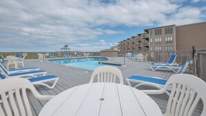 Un espace aménagé au bord de la piscine, avec des chaises longues et une table, une terrasse en bois et, au fond, un bâtiment doté de plusieurs balcons.