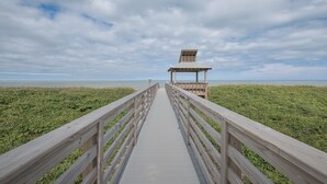 A wooden boardwalk leading to a small observation tower with a slanted roof, surrounded by a grassy area and the ocean in the background.