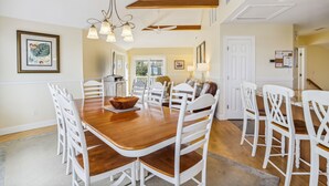 Un vaste espace dédié à la salle à manger, avec une table en bois et des chaises blanches, un lustre, et une vue sur la cuisine et le salon.