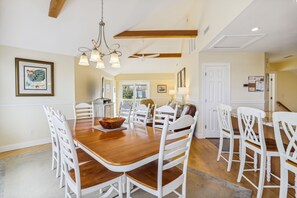 A spacious dining area with a wooden table and white chairs, a chandelier, and a view of the kitchen and living area. - 3675 - Seaducktion - Duck Getaway | Private Pool & Sleeps 10 (Duck)