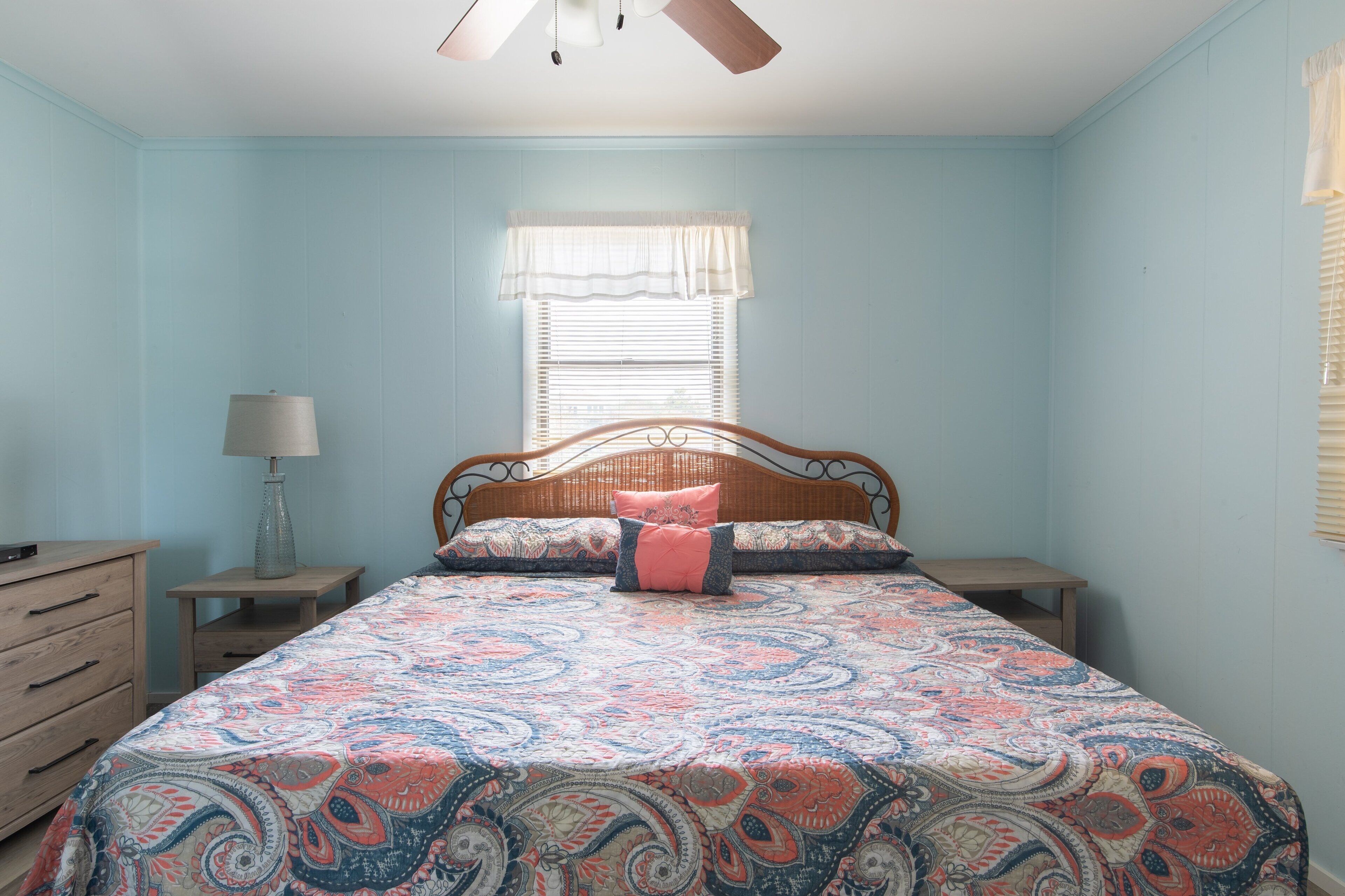 A bedroom with a bed, a nightstand, a dresser, a window with blinds, and a ceiling fan.