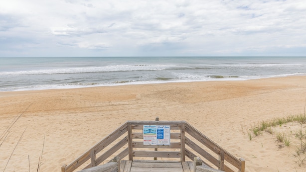 Ein Holzsteg führt zu einem Strand mit einem Hinweisschild.