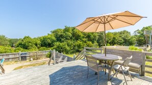 A wooden deck with a table and chairs, an umbrella, and a view of greenery. - 4601-Sandy Bluff - Southern Shores Home w/ Private Pool (Southern Shores)