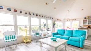 A bright living room with a turquoise sofa, wicker chairs, a white coffee table, and a kitchen area visible through large windows. - 4751- Purple Passion - Walk to Beach | Pool, Hot Tub & Dogs Welcome (Kitty Hawk)