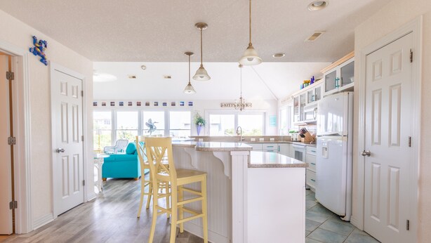 A modern kitchen with white cabinetry, a central island with bar stools, pendant lighting, and a view to a bright living area. - 4751- Purple Passion - Walk to Beach | Pool, Hot Tub & Dogs Welcome (Kitty Hawk)