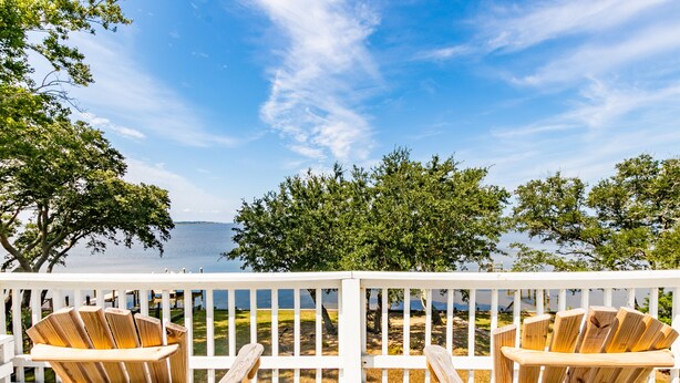A wooden deck with two Adirondack chairs facing a body of water, surrounded by trees and a white railing. - 5002 - Bohannon - Soundfront KDH Escape w/ Sunset Views (Kill Devil Hills)
