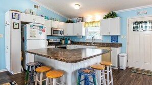 A kitchen with a central island, white cabinets, a microwave, and a sink. There are three stools around the island. - 5002 - Bohannon - Soundfront KDH Escape w/ Sunset Views (Kill Devil Hills)