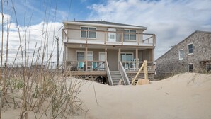 A two-story beach house with a balcony, wooden deck, and a sandy beach in the background. - 5061 - Good Night Irene - Oceanfront Cottage w/ Decks & Stunning Views (Kill Devil Hills)