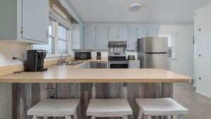 A modern kitchen with light blue cabinets, a wooden island with bar stools, stainless steel appliances, and a window with blinds. - 5061 - Good Night Irene - Oceanfront Cottage w/ Decks & Stunning Views (Kill Devil Hills)