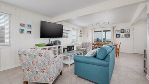 A living room with a television, patterned armchair, sofa, and dining area with a view. - 5061 - Good Night Irene - Oceanfront Cottage w/ Decks & Stunning Views (Kill Devil Hills)